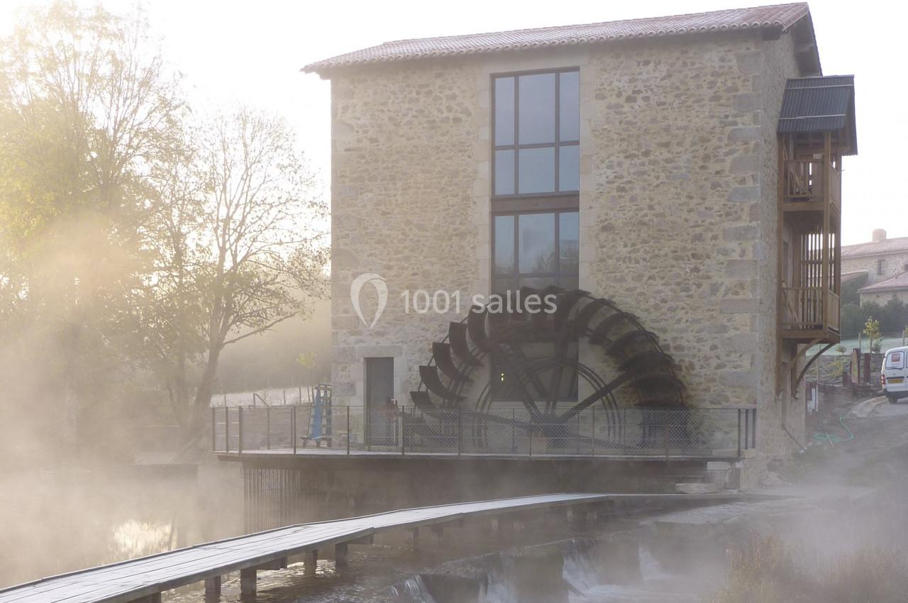 Bâtiment en pierre avec une grande roue à aubes, entouré de brume et bordé d'un cours d'eau.