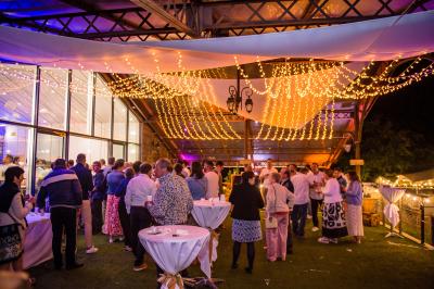 Salle de réception éclairée en rose avec un buffet dressé et des tables hautes décorées de petites lampes.