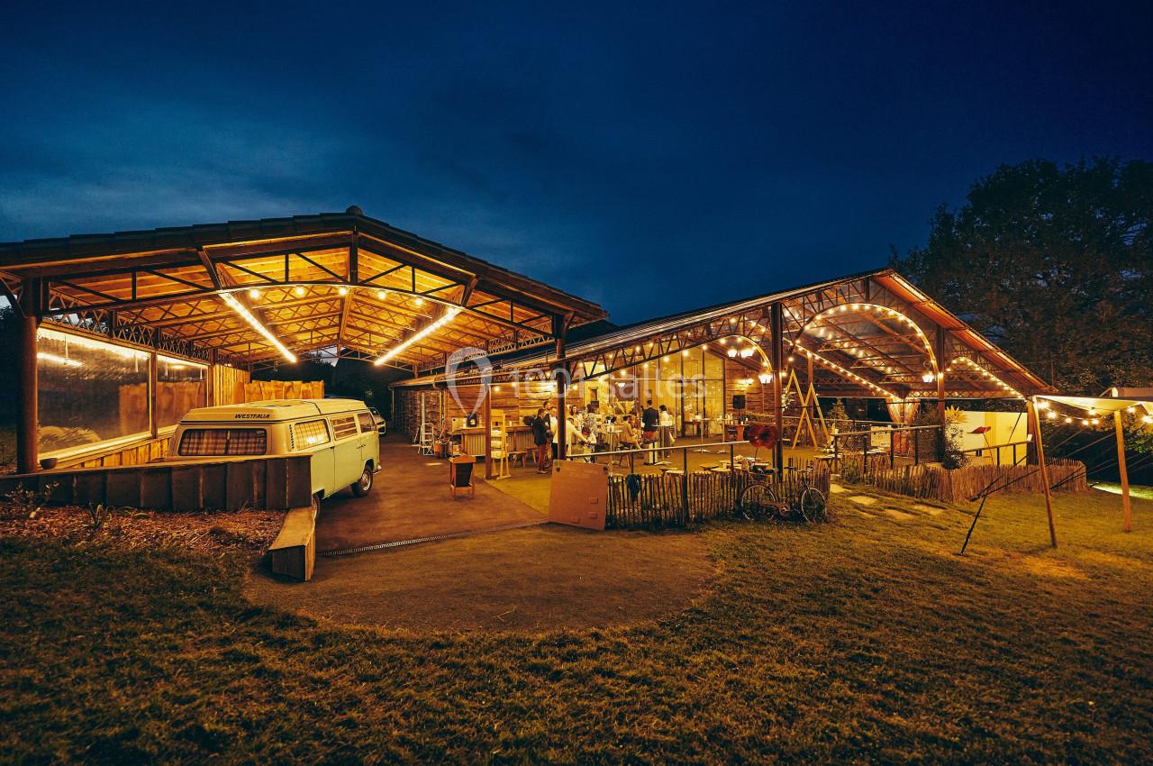 Espace extérieur éclairé avec des guirlandes lumineuses, abritant une structure en bois et un van stationné.