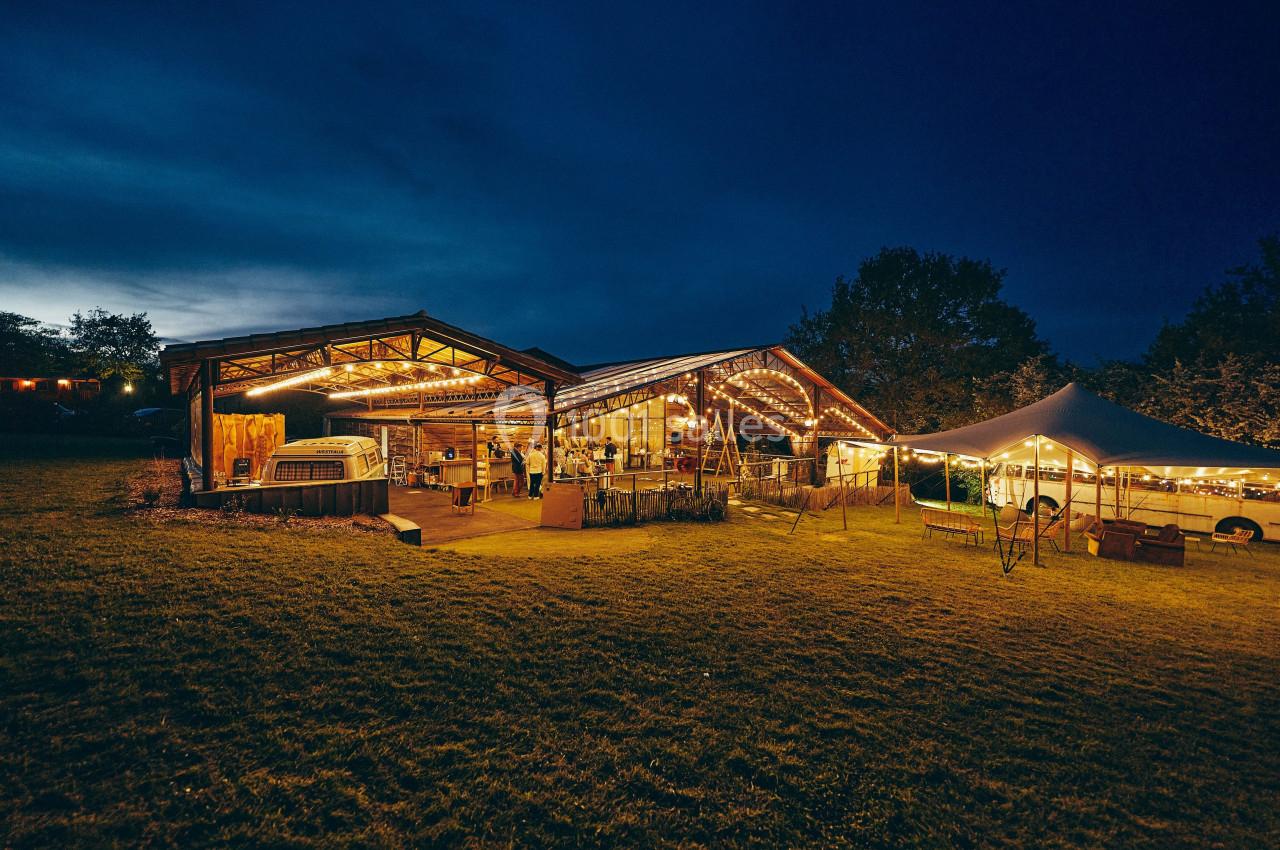 Grange éclairée en soirée avec des guirlandes lumineuses, entourée d'une pelouse et d'une tente blanche.