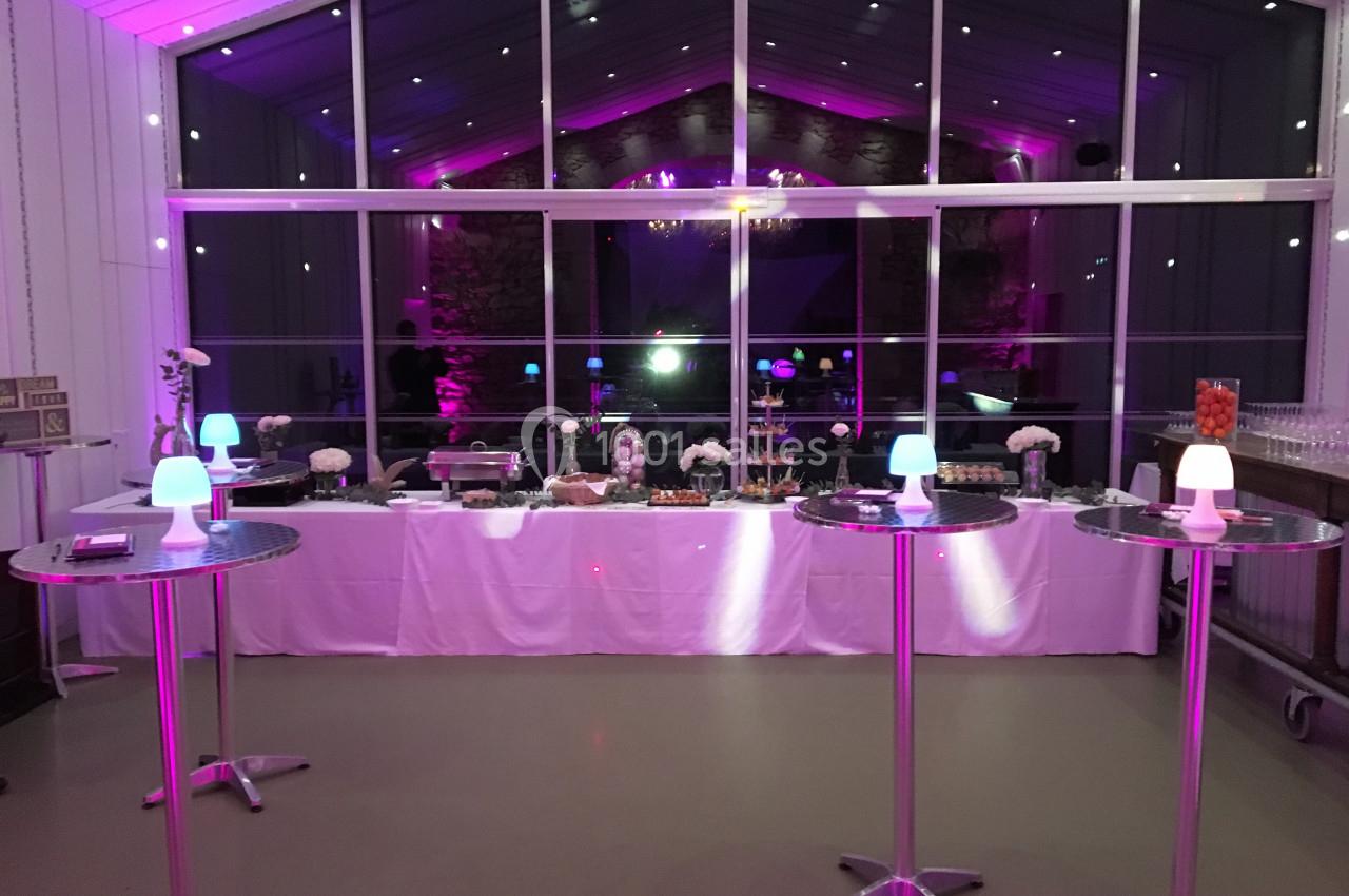 Salle de réception éclairée en rose avec un buffet dressé et des tables hautes décorées de petites lampes.
