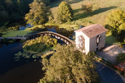 Bâtiment en pierre au bord d'une rivière entourée de végétation, avec un pont en arrière-plan sous un ciel dégagé.