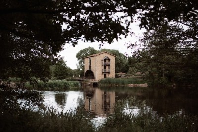 Bâtiment en pierre au bord d'une rivière entourée de végétation, avec un pont en arrière-plan sous un ciel dégagé.