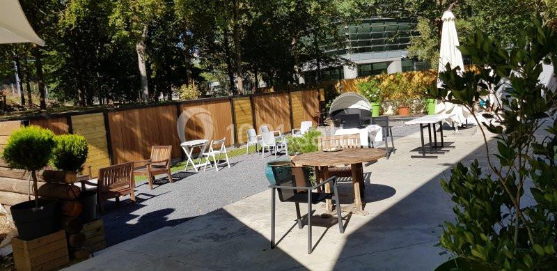 Terrasse extérieure avec tables, chaises, parasols et végétation, entourée de clôtures en bois et d'arbres.