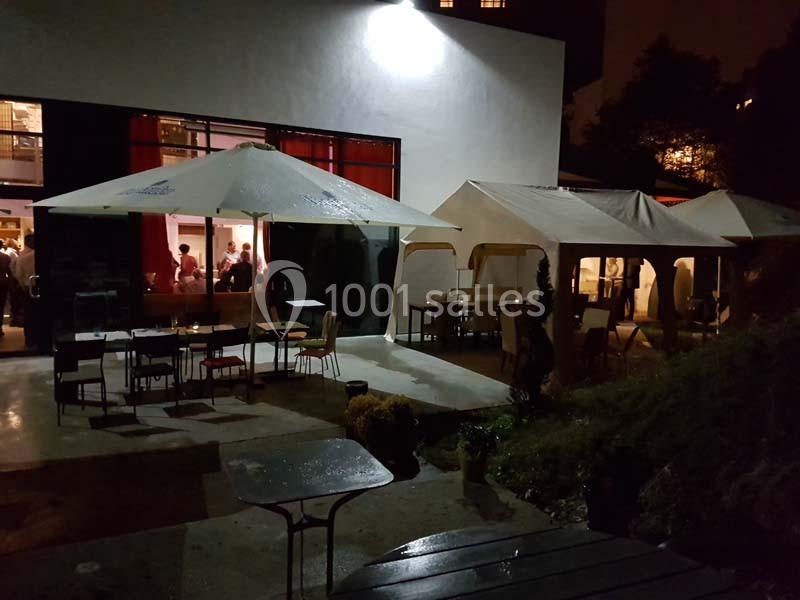 Terrasse extérieure avec tables et chaises sous des tonnelles, éclairée la nuit, avec des personnes visibles à l'intérieur.