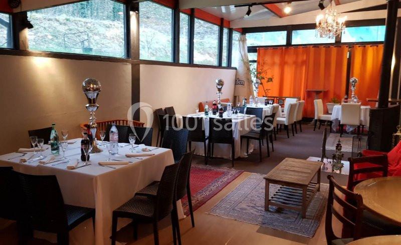 Salle de restaurant lumineuse avec tables dressées, nappes blanches, chaises noires et rideaux orange en arrière-plan.