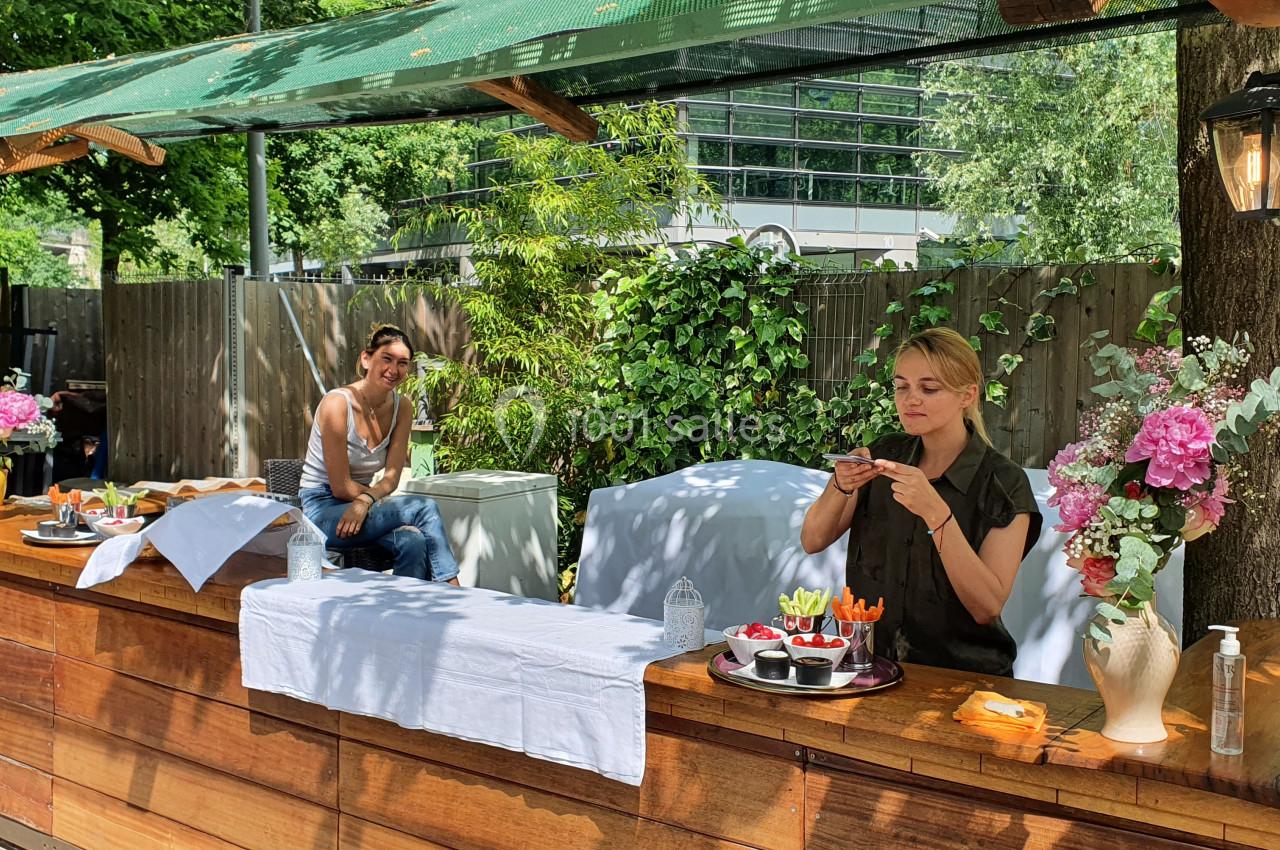 Deux femmes dans un stand en bois en extérieur, l'une préparant des plats, l'autre assise en arrière-plan.