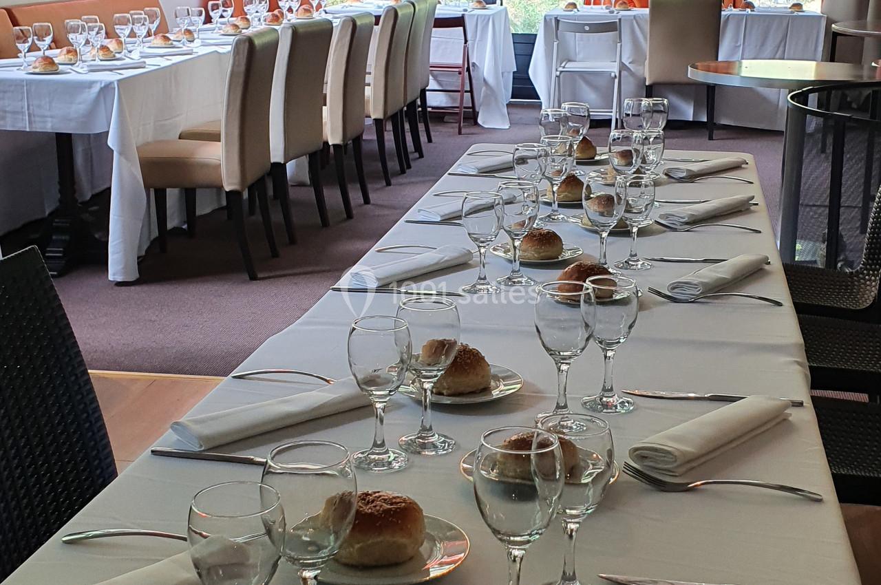 Salle de réception lumineuse avec tables dressées, nappes blanches, verres, assiettes et petits pains alignés.
