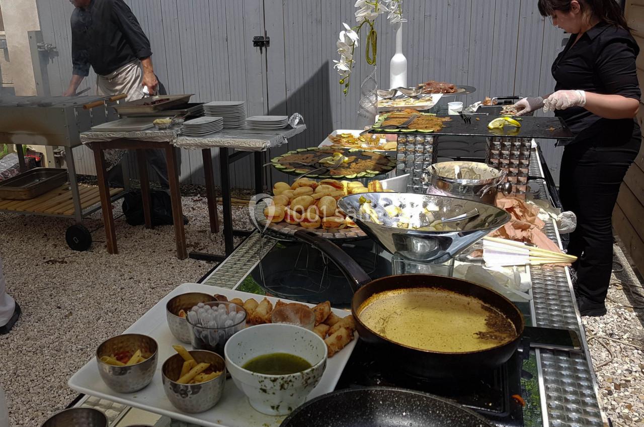 Buffet en extérieur avec plats variés, cuisinier au barbecue et serveuse préparant des assiettes.