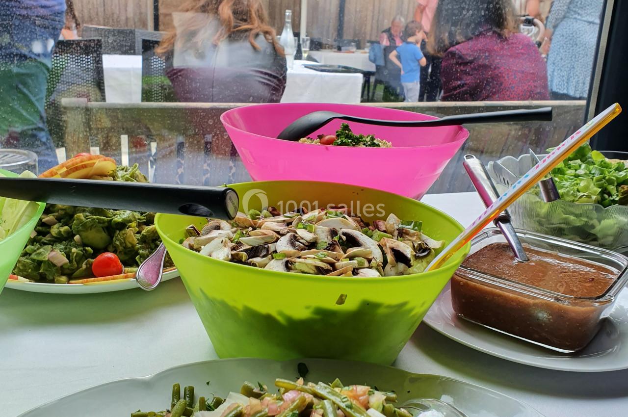 Buffet avec salades variées en premier plan, terrasse extérieure avec convives en arrière-plan sous des parasols.