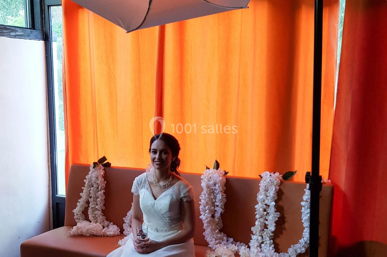 Une femme en robe de mariée est assise sur un canapé, éclairée par un parapluie de studio devant des rideaux orange.