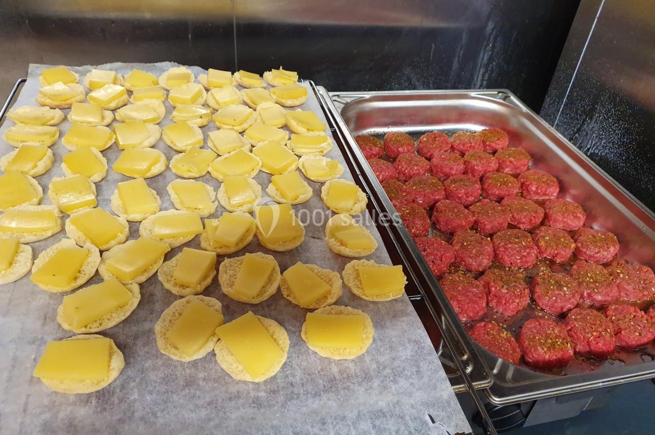 Petits pains garnis de fromage à gauche et mini-steaks hachés crus alignés dans un plat à droite, en cuisine.