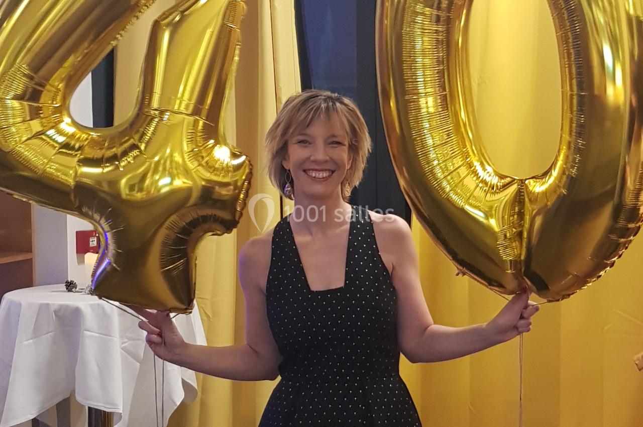 Femme souriante tenant des ballons dorés formant le chiffre 40 dans une salle décorée de rideaux jaunes.