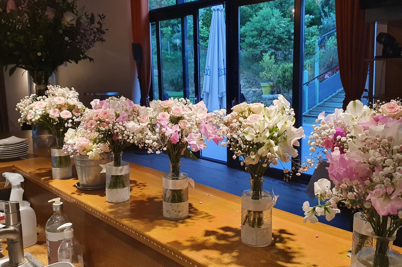 Bouquets de fleurs roses et blanches alignés sur un comptoir en bois, avec une grande baie vitrée en arrière-plan.