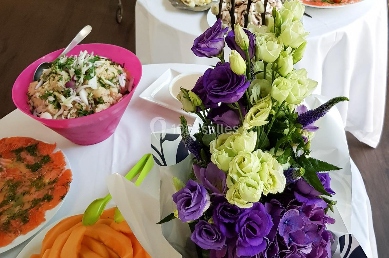 Buffet avec des plats variés, dont des salades, du saumon fumé et des fruits, décoré d'un bouquet de fleurs violettes et…