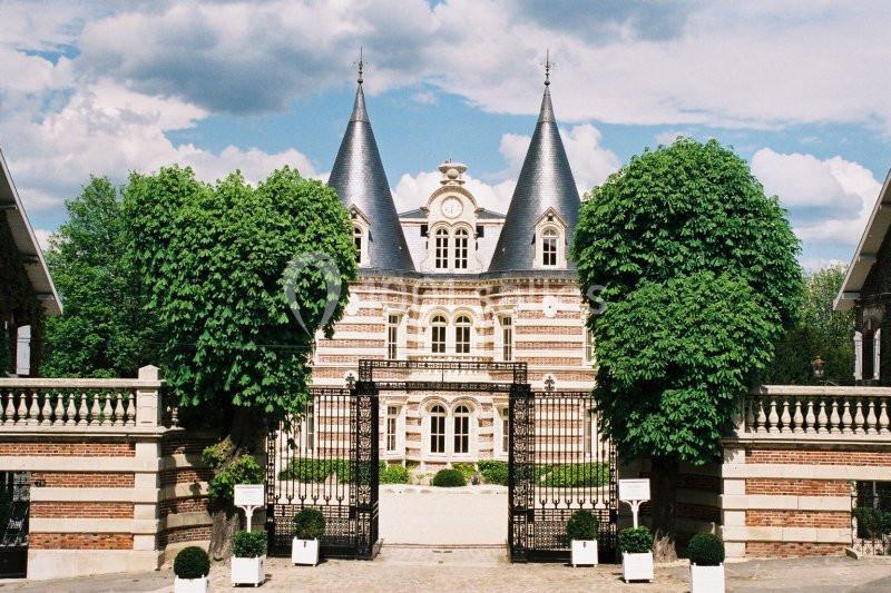 Façade d'un château en briques avec deux tours, entouré d'arbres et d'une grille en fer forgé.
