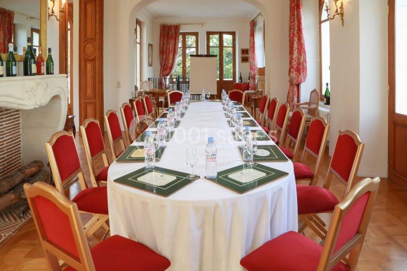 Salle de réunion lumineuse avec une grande table ovale dressée, entourée de chaises rouges et décorée de bouteilles de vin.