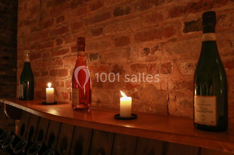 Trois bouteilles de vin et trois bougies allumées posées sur une étagère en bois devant un mur de briques.