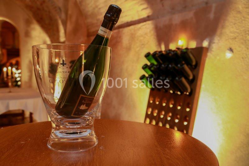 Bouteille de vin pétillant dans un seau transparent sur une table en bois, avec un porte-bouteilles en arrière-plan.