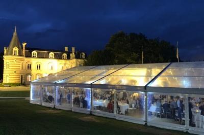 Vue nocturne d'une réception dans une tente éclairée, installée devant un bâtiment de style château.