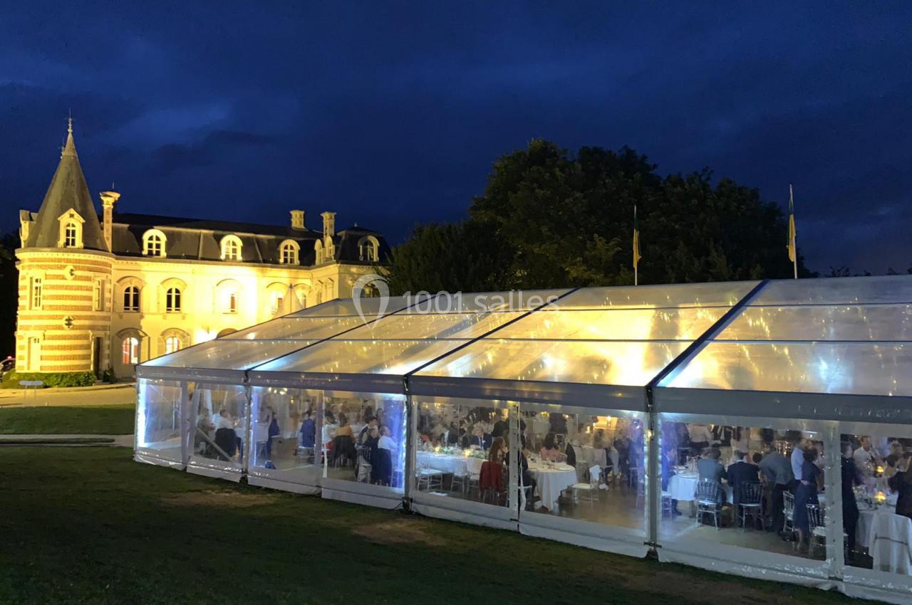 Vue nocturne d'une réception dans une tente éclairée, installée devant un bâtiment de style château.