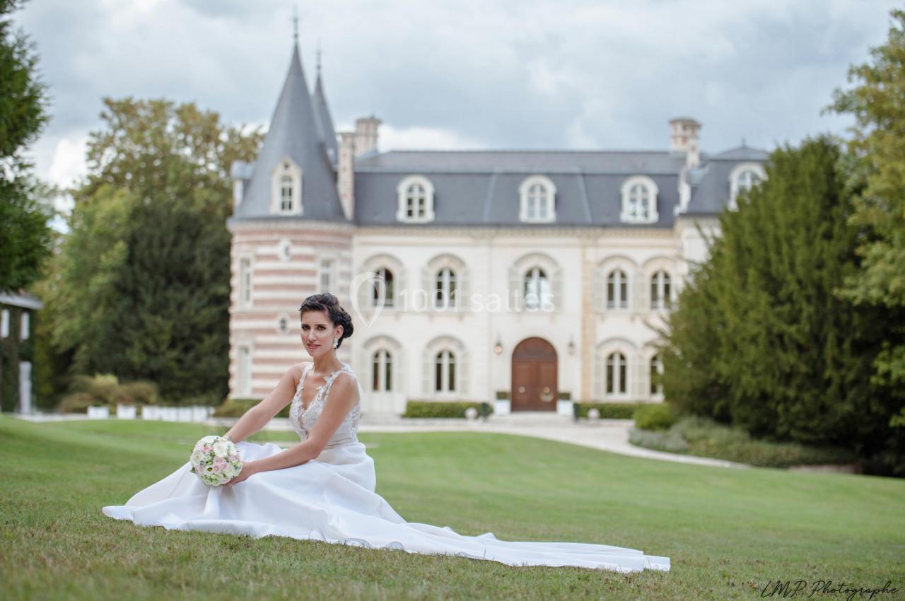 Une mariée en robe blanche est assise sur une pelouse devant un château au style classique.