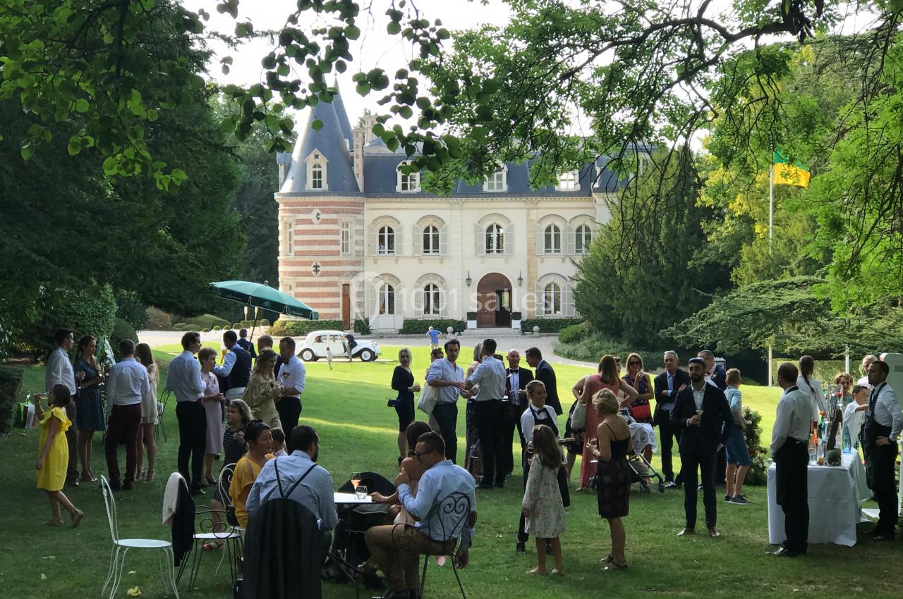 Groupe de personnes rassemblées dans un jardin devant un bâtiment de style château, lors d'un événement en plein air.