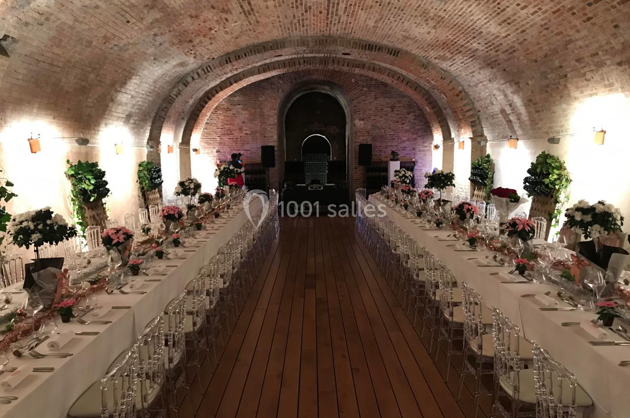 Salle voûtée en briques avec deux longues tables dressées pour un événement, décorée de fleurs et éclairée chaleureusement.