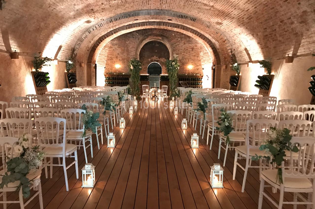 Salle voûtée décorée pour une cérémonie, avec allée centrale bordée de lanternes et chaises blanches alignées.