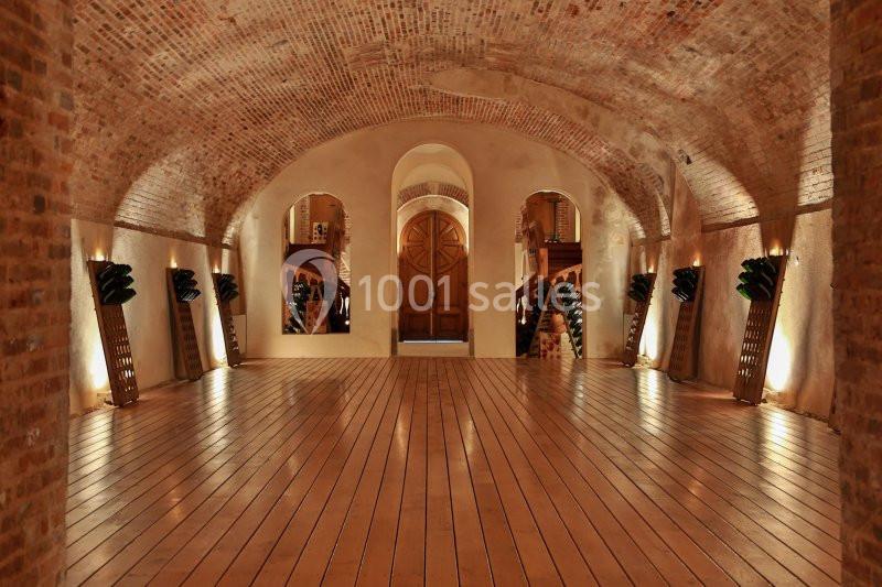 Salle voûtée en briques avec un sol en bois, éclairage tamisé et porte en bois au fond.