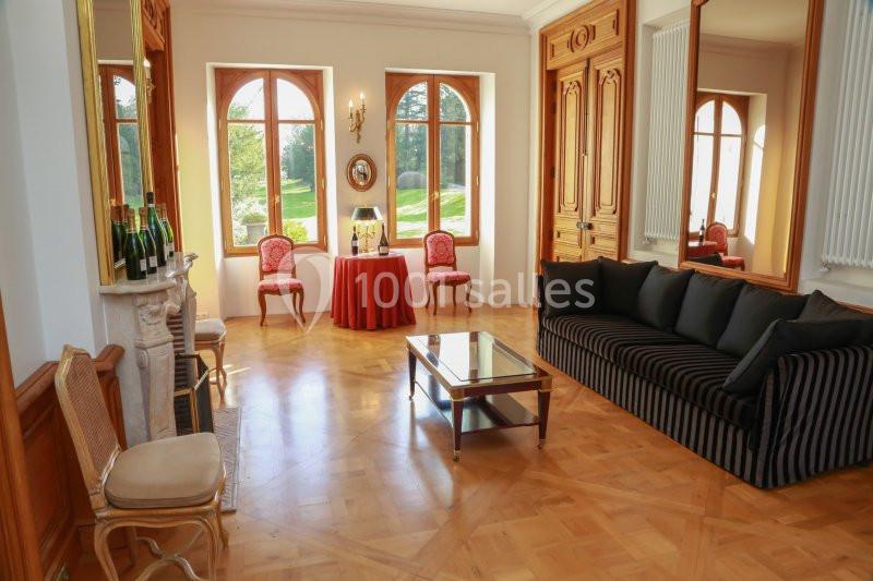 Salon lumineux avec parquet, canapé noir, chaises classiques, cheminée, table basse et grandes fenêtres donnant sur un…