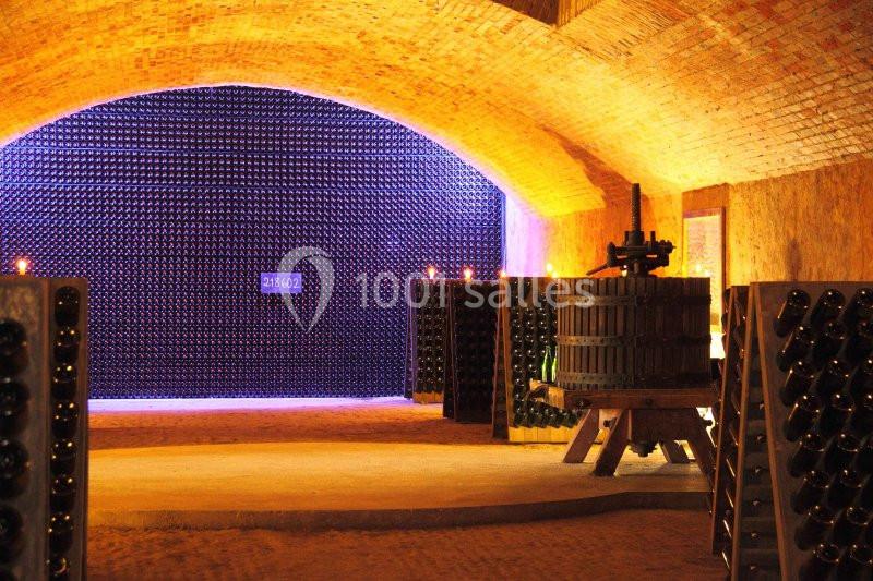 Cave voûtée avec des murs en briques, des bouteilles de vin empilées et une ancienne presse en bois.