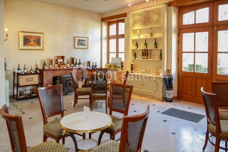 Salle lumineuse avec des tables et chaises en bois, étagères exposant des bouteilles et fenêtres donnant sur l'extérieur.
