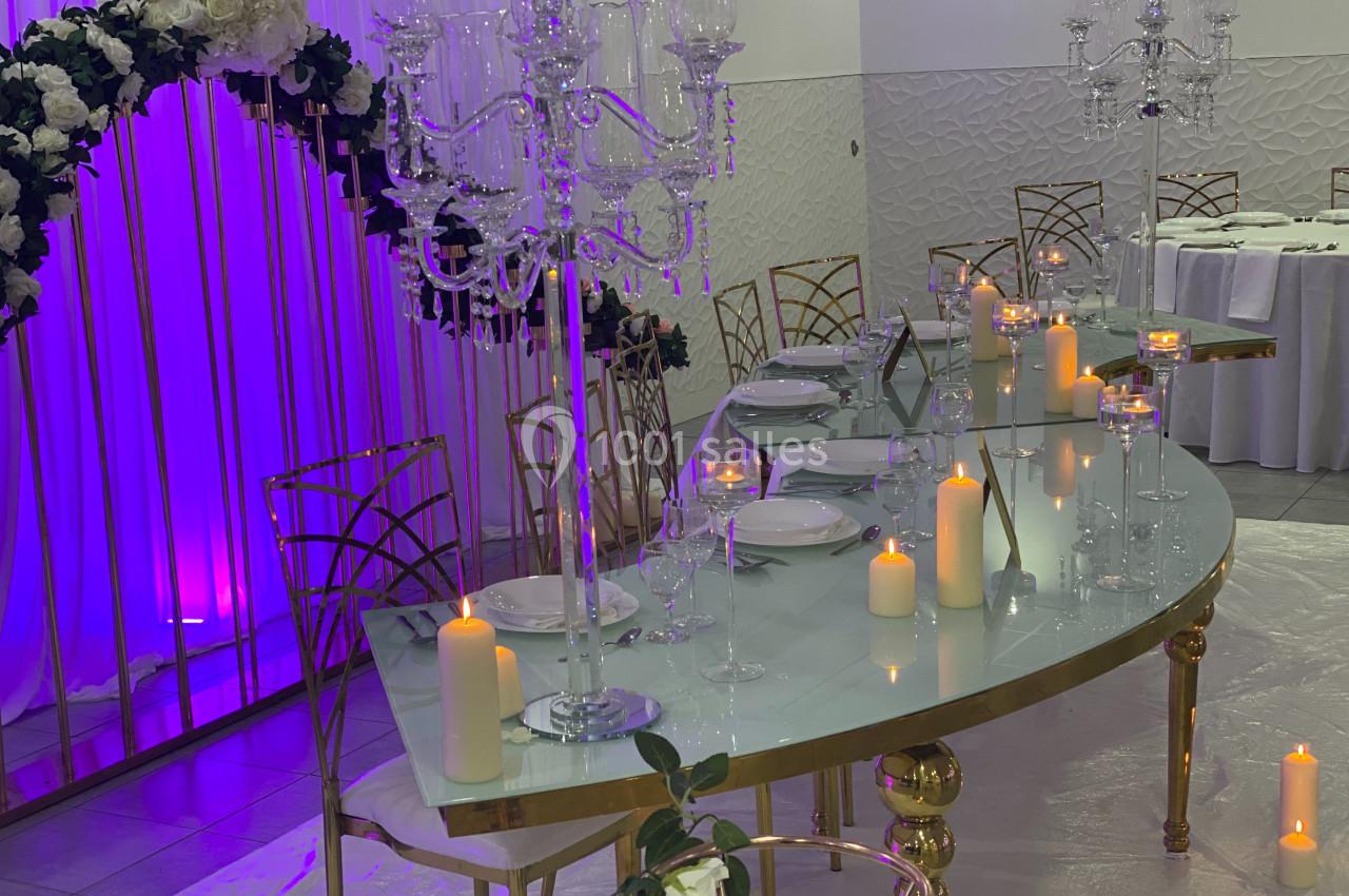 Table élégante décorée pour un événement, avec chandeliers, bougies, vaisselle blanche et fond lumineux violet.