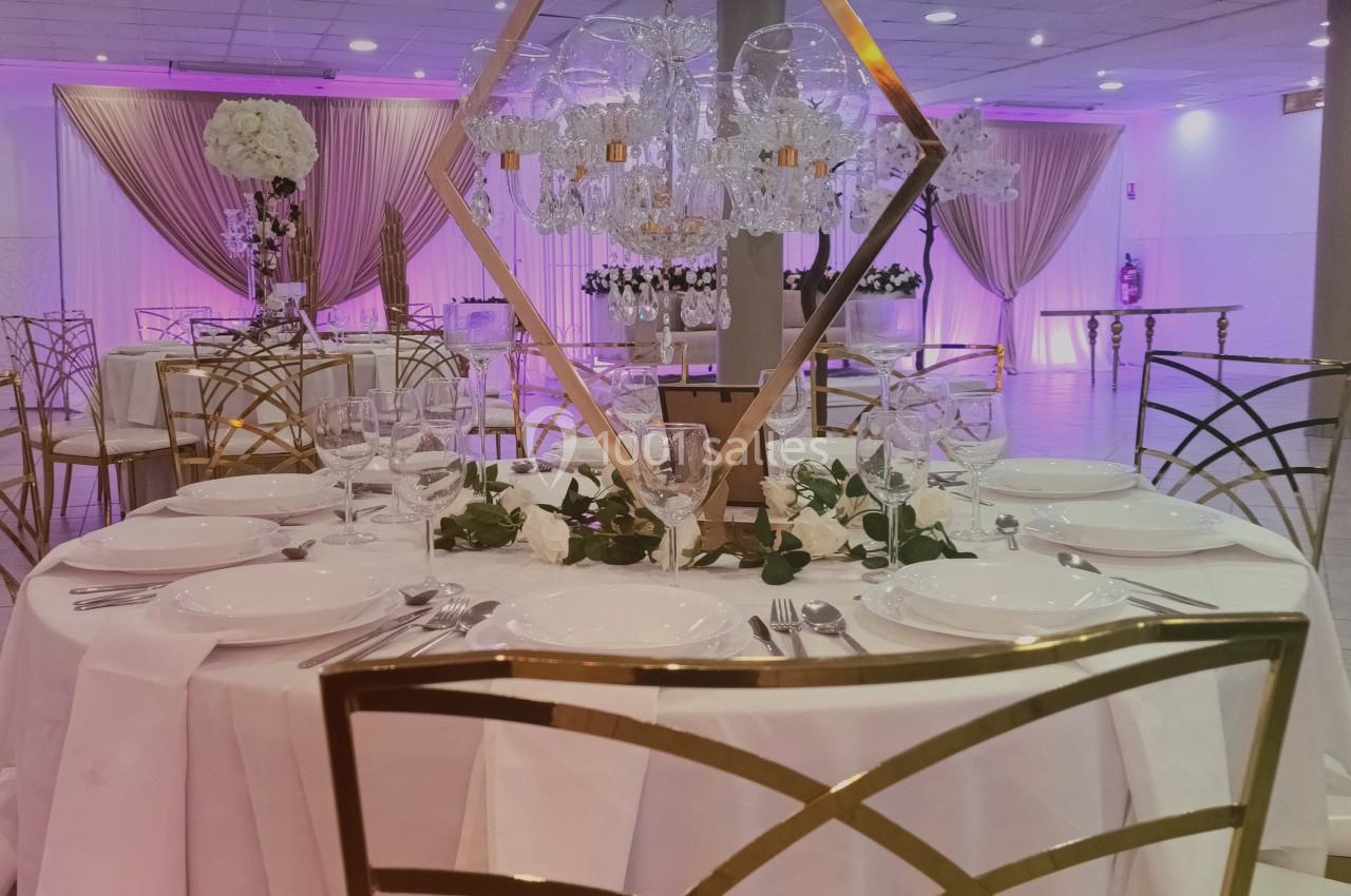 Salle de réception décorée avec des tables élégantes, chaises dorées, nappes blanches et éclairage rose tamisé.