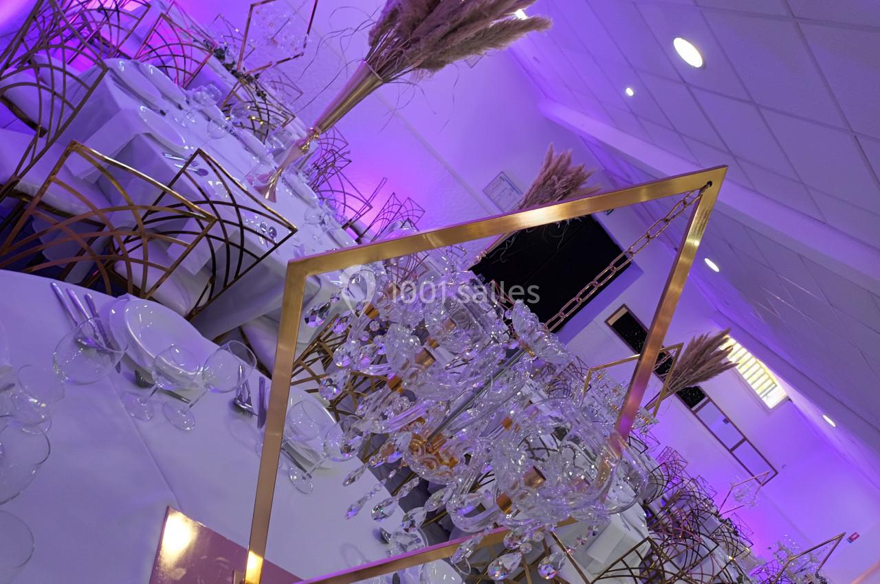 Salle de réception décorée avec des tables dressées, des chaises dorées et des ornements floraux sous éclairage violet.