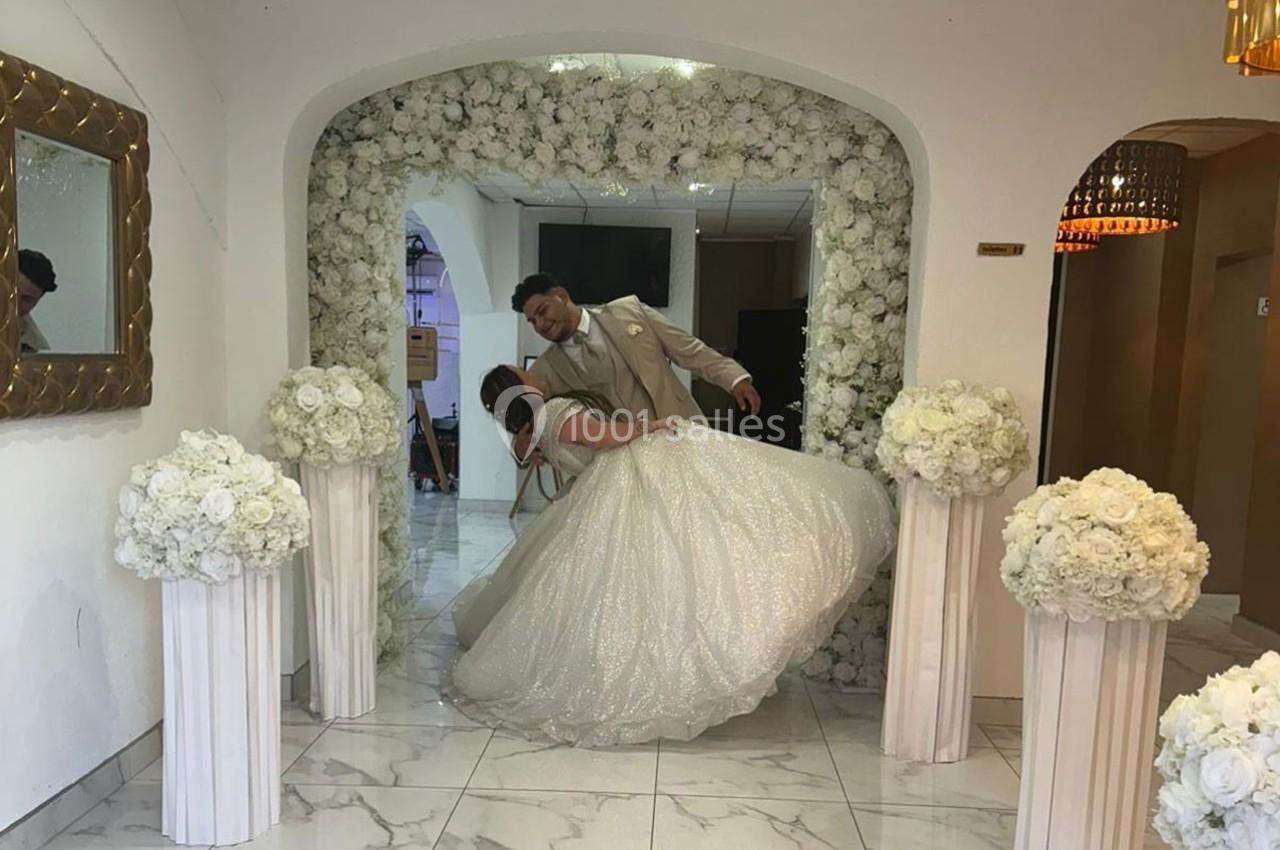 Un couple en tenue de mariage danse sous une arche décorée de fleurs blanches dans une salle lumineuse.