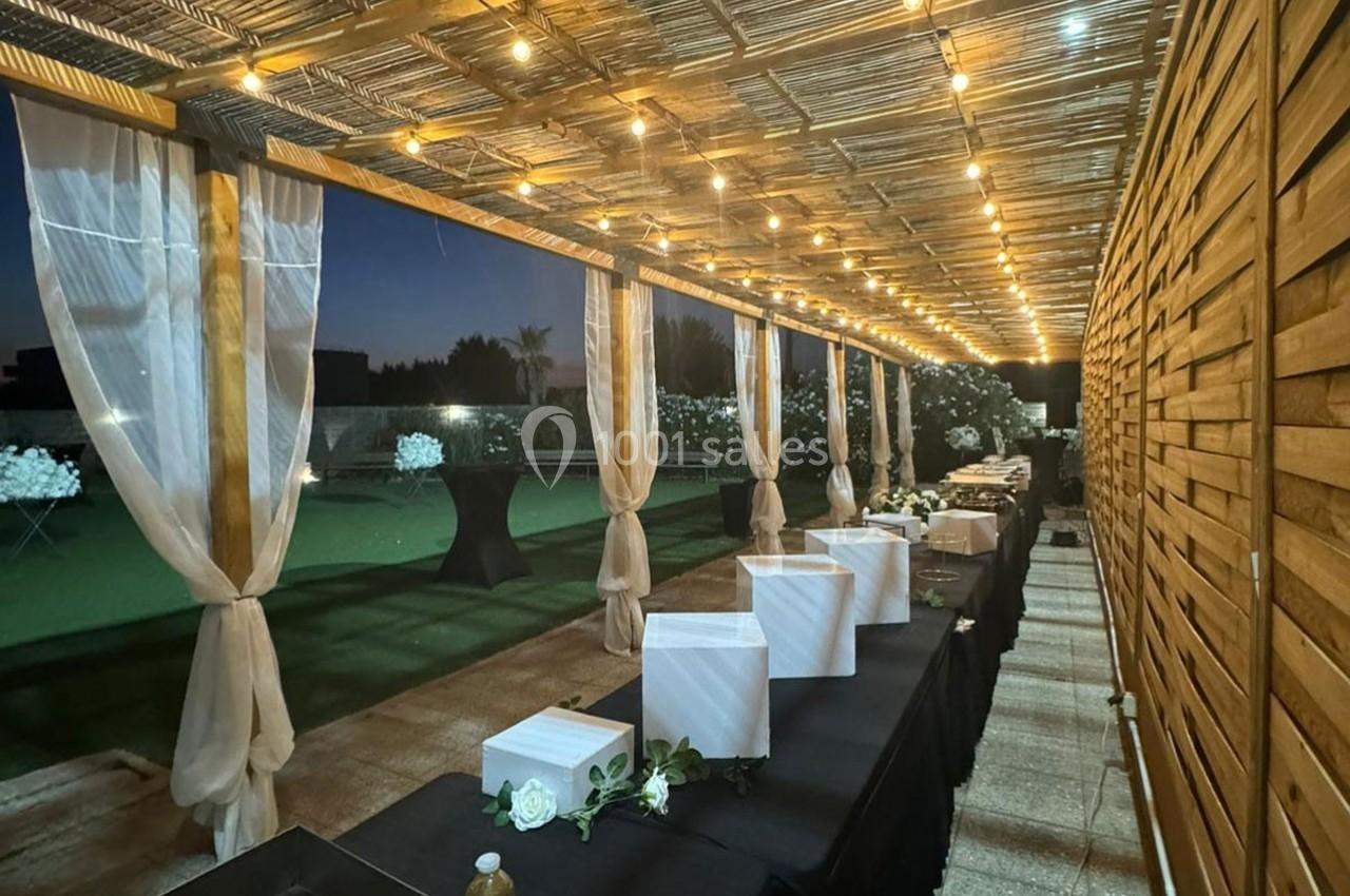 Allée couverte éclairée par des guirlandes lumineuses, avec des tables dressées et un jardin en arrière-plan.