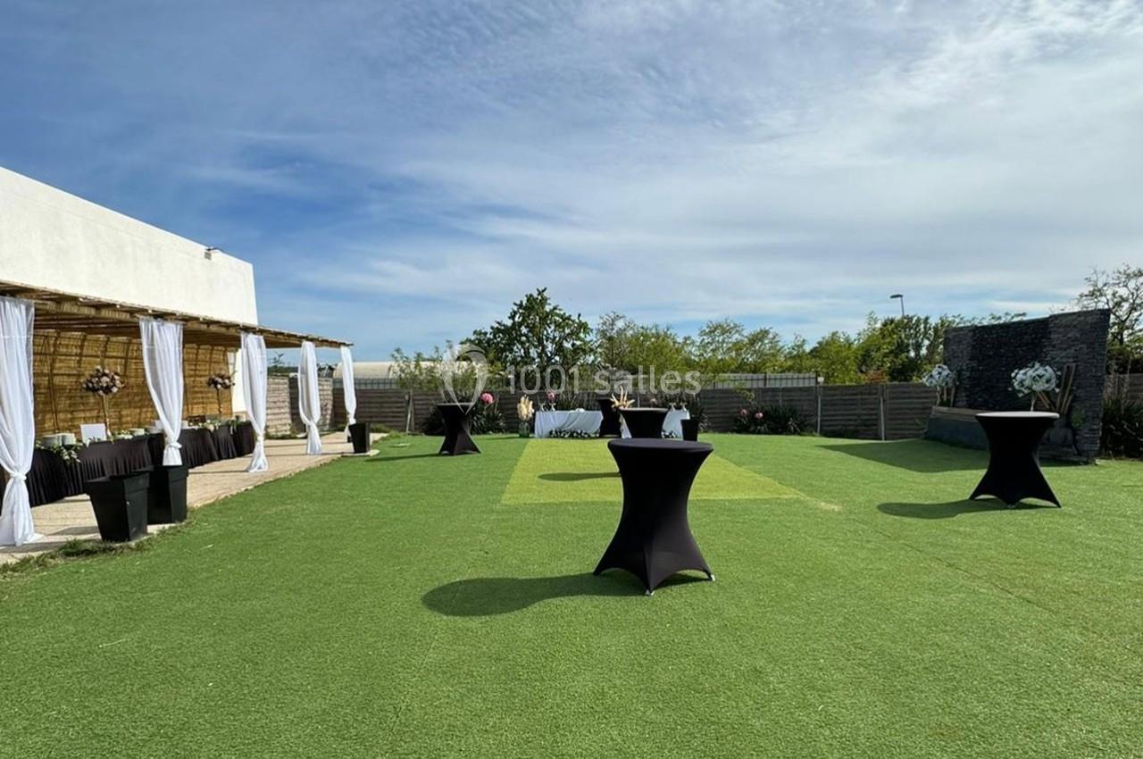 Jardin aménagé avec pelouse artificielle, tables hautes noires et espace décoré pour un événement en plein air.