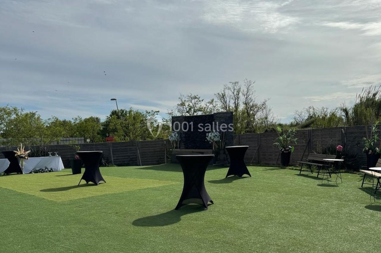 Espace extérieur avec pelouse synthétique, tables hautes noires et quelques plantes en pot, sous un ciel partiellement…