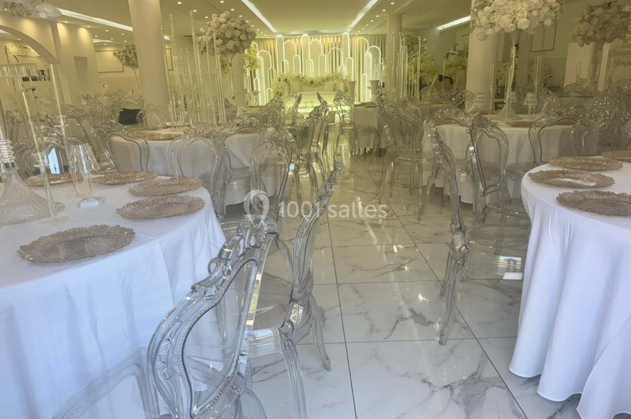 Salle de réception élégante avec tables rondes dressées, chaises transparentes et décorations florales blanches.