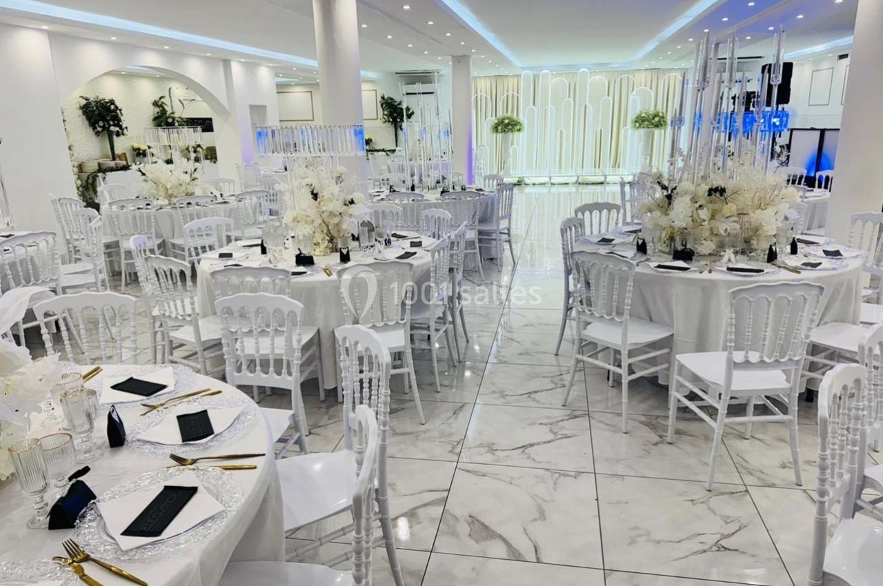 Salle de réception élégante avec tables rondes décorées de nappes blanches, fleurs et couverts dorés.