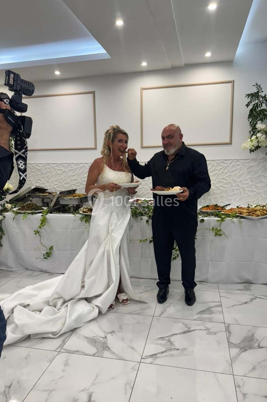 Une femme en robe blanche et un homme en noir mangent devant un buffet décoré de fleurs dans une salle lumineuse.