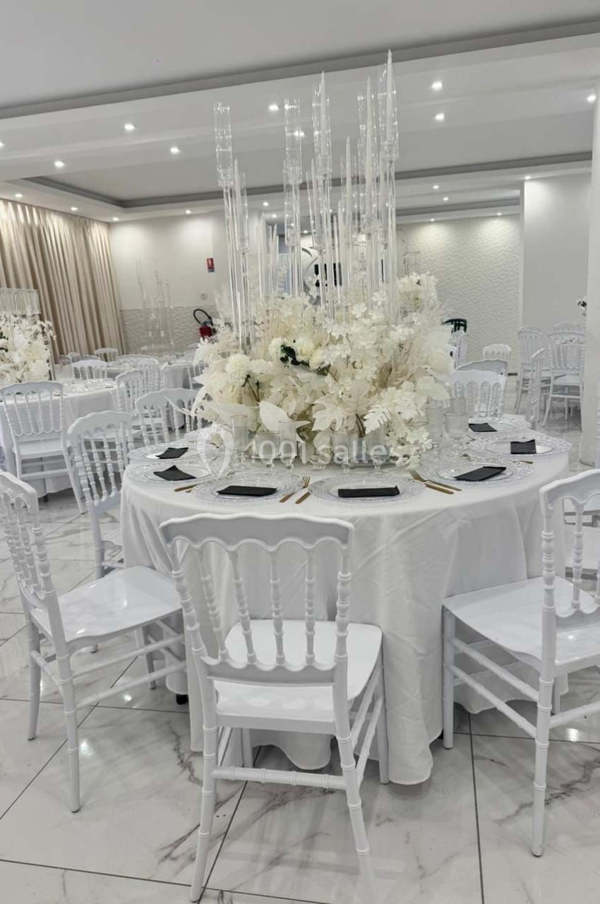Salle de réception décorée avec des tables rondes, nappes blanches, chaises blanches et centre de table floral élégant.