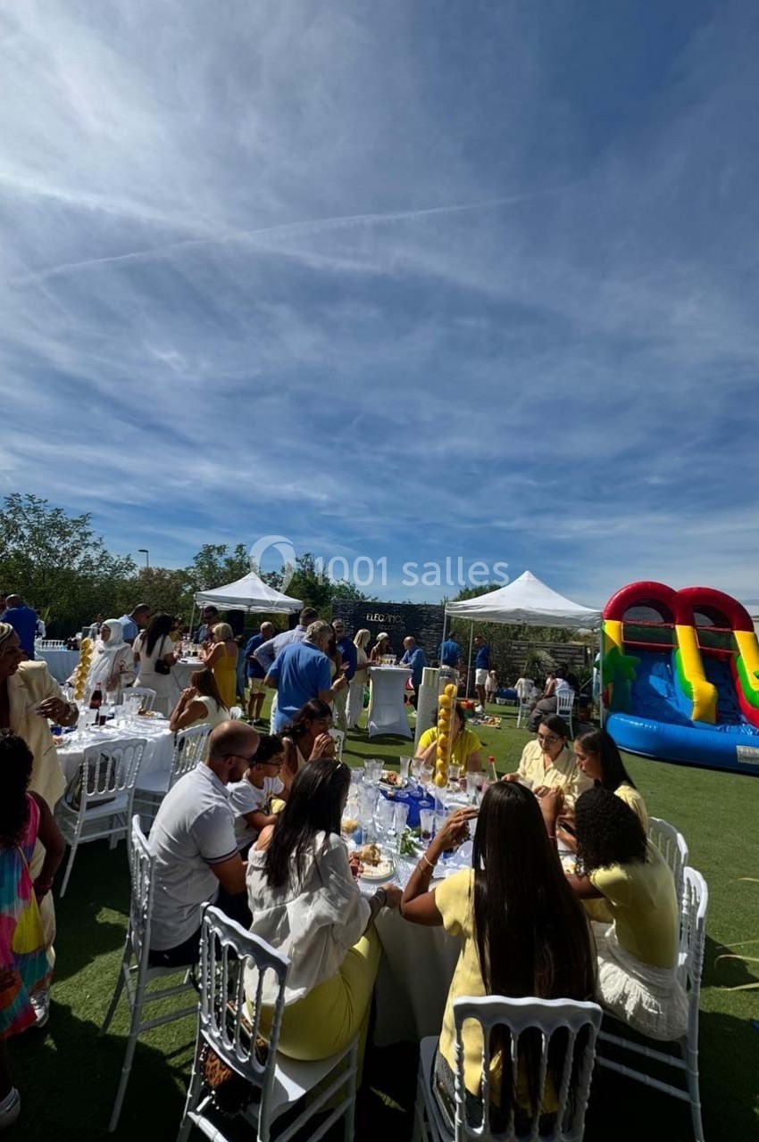 Des personnes assises et debout lors d'un événement en plein air avec des tables, des tentes blanches et un château…