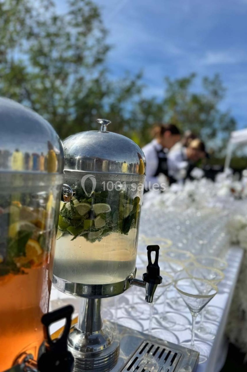 Distributeurs de boissons infusées avec fruits et herbes sur une table en extérieur, avec des verres alignés en arrière-plan.