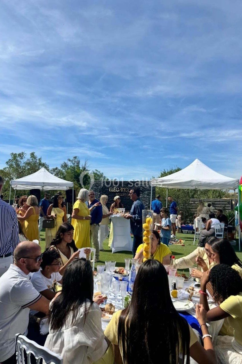 Des personnes rassemblées à l'extérieur lors d'un événement sous des tentes blanches, avec des tables dressées et un château…