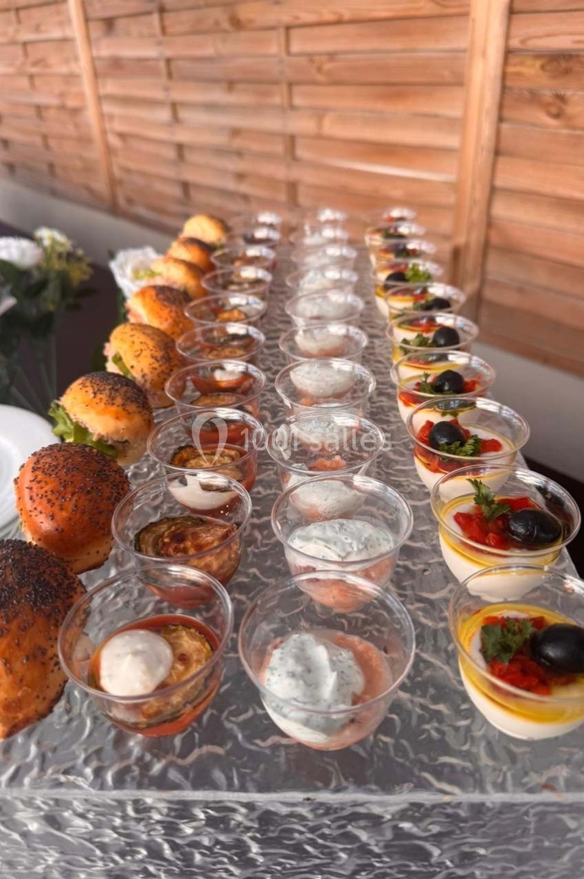 Plateau de verrines et mini-burgers disposés en rangées sur une table en verre, devant un fond en bois.