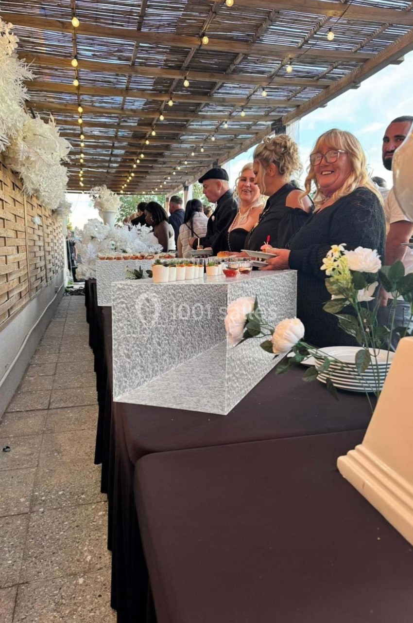 Des personnes souriantes debout derrière une table de buffet décorée de fleurs blanches sous une pergola éclairée.