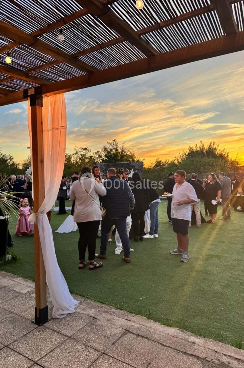 Personnes rassemblées dans un jardin au coucher du soleil, sous une pergola en bois décorée de voilages blancs.